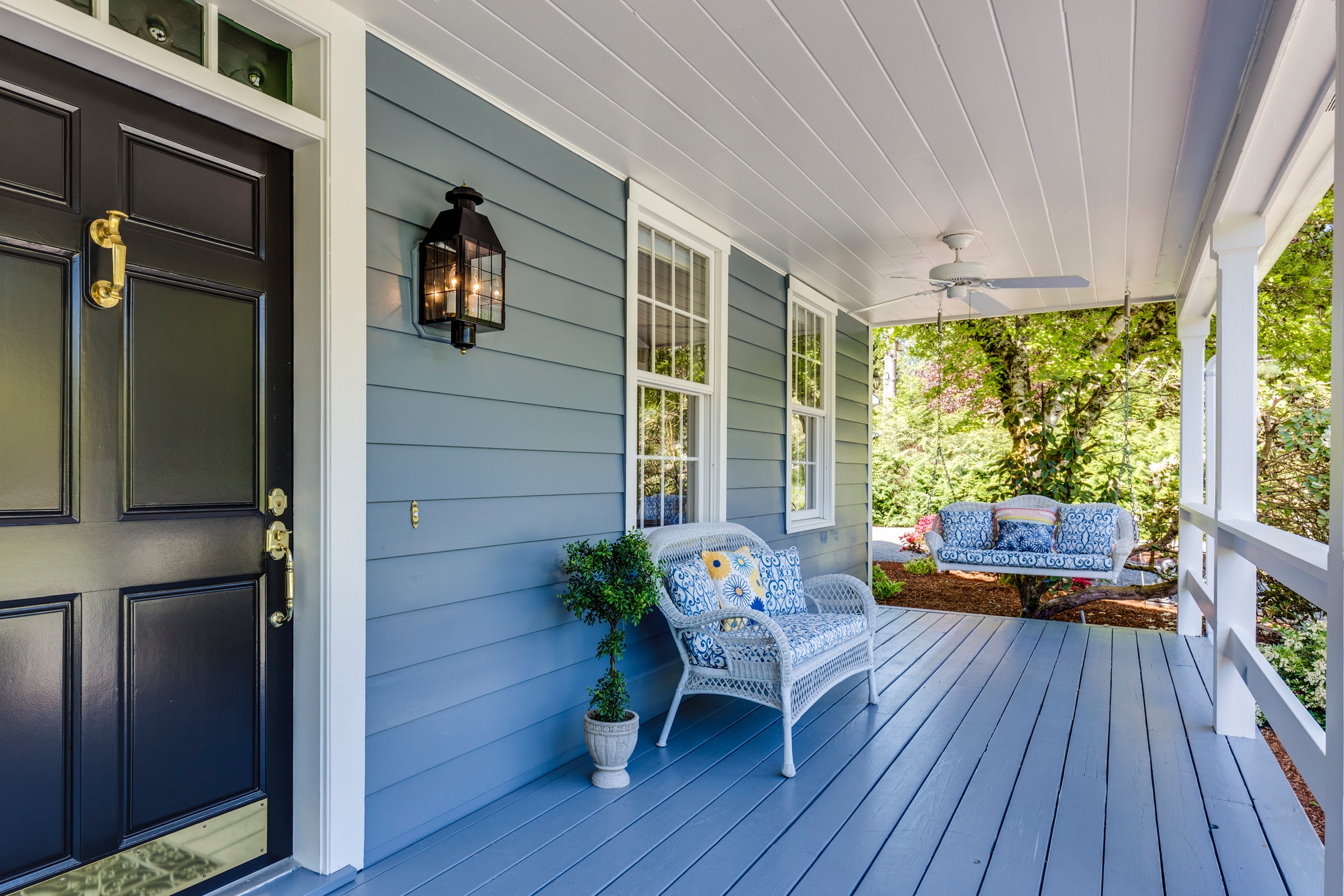 Blue house front porch with a black door, and a whicker porch swing.