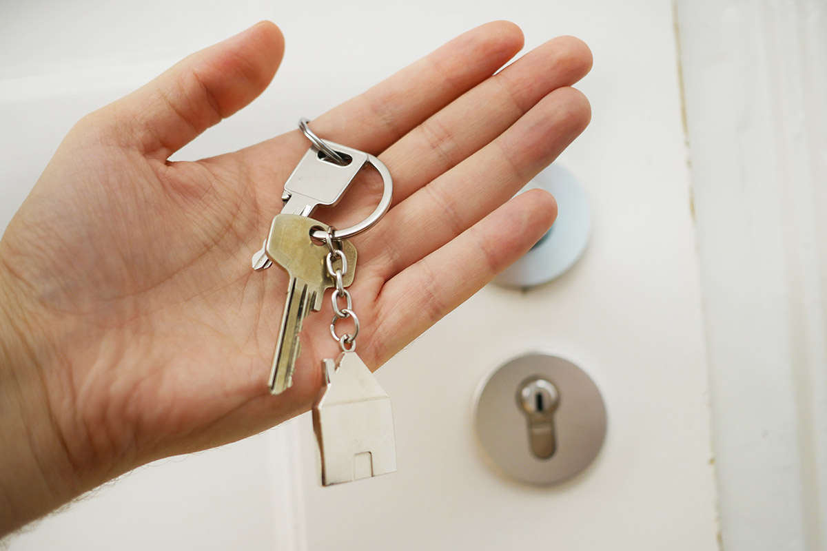 hand holding keys on keychain with small metal house and door in the background