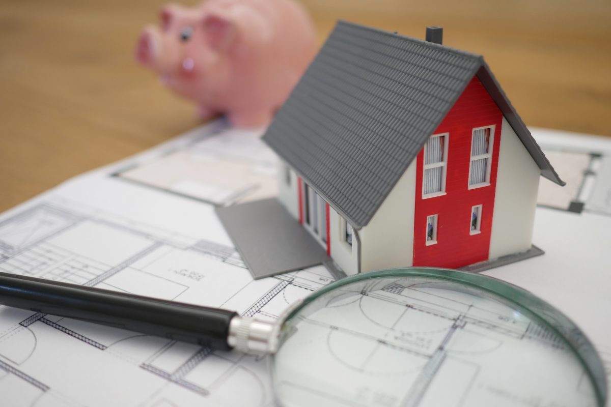 Magnifying glass and miniature house sitting on house blueprints with blurry piggy bank in background
