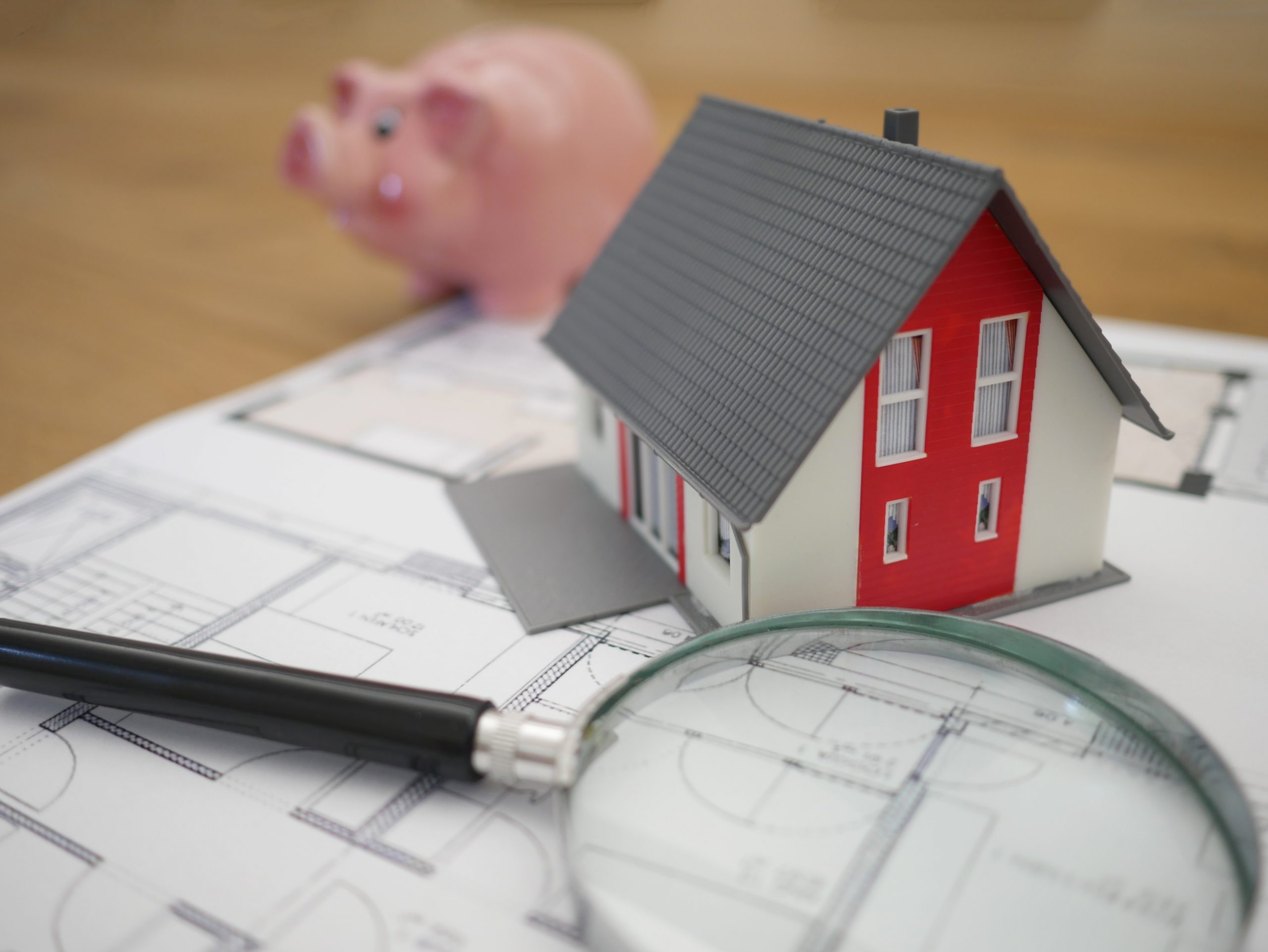 Magnifying glass and miniature house sitting on house blueprints with blurry piggy bank in background